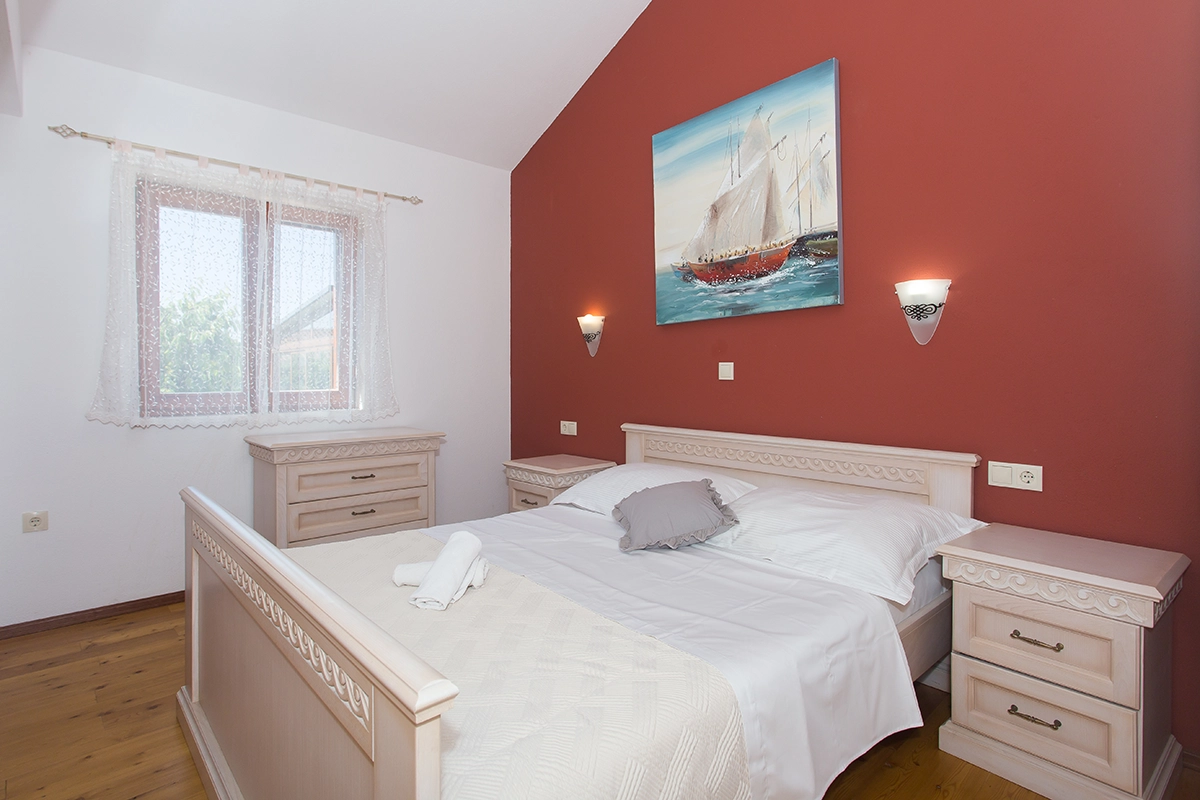 Room with a double bed, one red wall and one white wall, a window, and nightstands on each side of the bed.